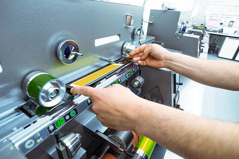Hände justieren einen Papierstreifen auf einer Andruckmaschine zur Qualitätskontrolle einer Farbprobe.
