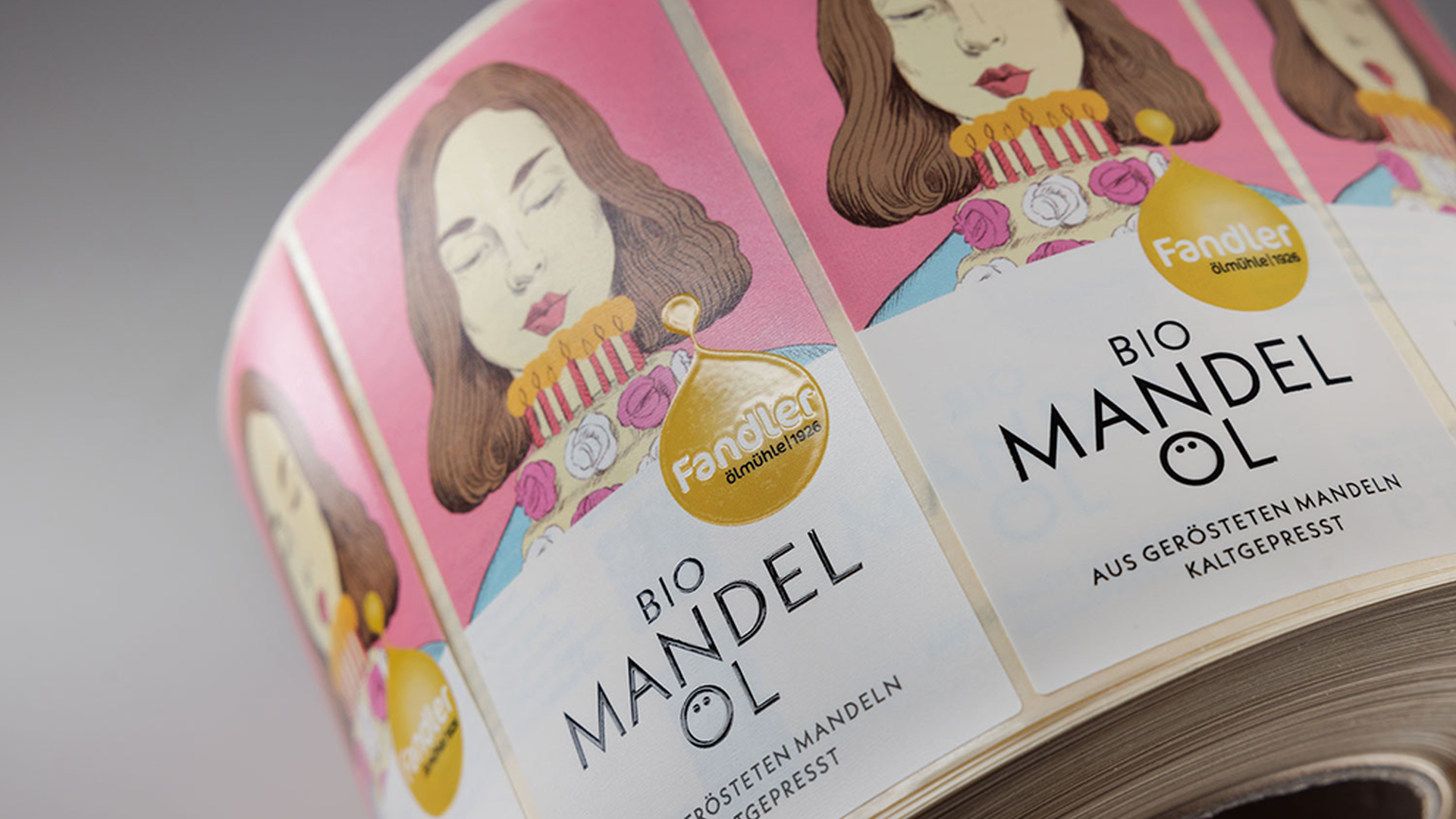 Rolle mit Etiketten „Bio Mandelöl“ in Rosa und Weiß mit goldfarbenem Logo und illustriertem Porträtmotiv.